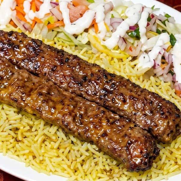 Two glistening grilled kebabs on a bed of yellow rice with a side salad and white sauce.